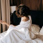 A woman sitting on an unmade bed in morning light, turned away, one hand outstretched.