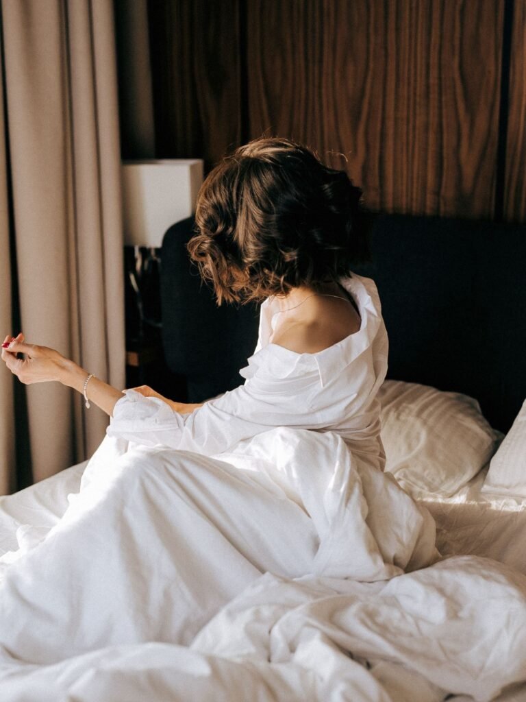 A woman sitting on an unmade bed in morning light, turned away, one hand outstretched.