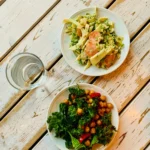 Two plates of different food on a weathered white wooden table, seen from above, with a glass of water beside them.
