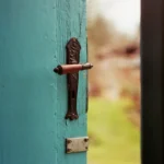 A teal wooden door standing ajar, the handle within reach, a green field visible through the opening.