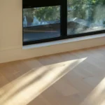 An empty room with a tall corner window, sunlight falling in long shapes across a bare wooden floor.